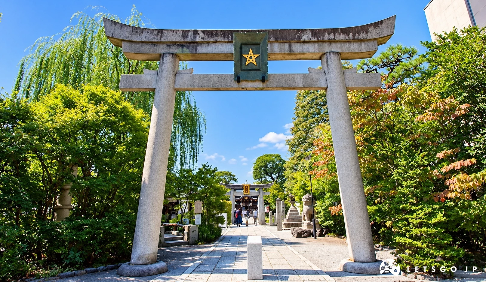 石造鳥居的中央匾額有金色五芒星標誌,參道兩旁有綠樹與垂柳,遠處有行人與第二座鳥居