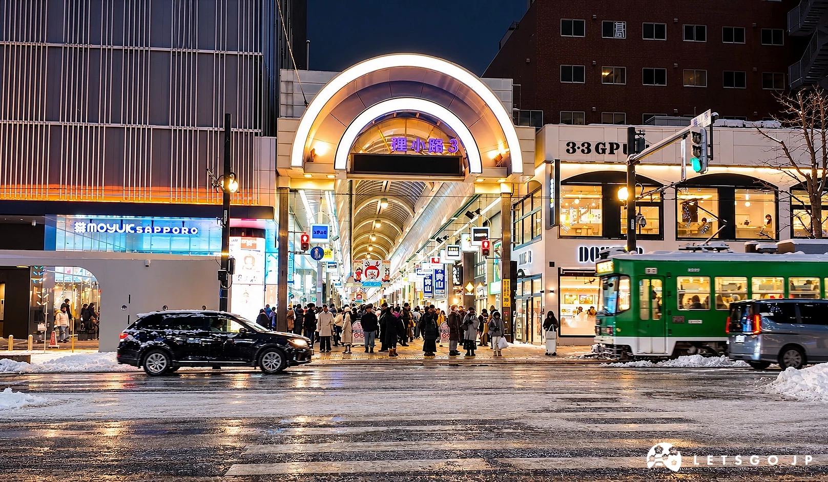 夜間濕滑的路口前有「狸小路3」拱廊入口與人群,右側有綠色路面電車,左側有「MOYUK SAPPORO」招牌與黑色汽車