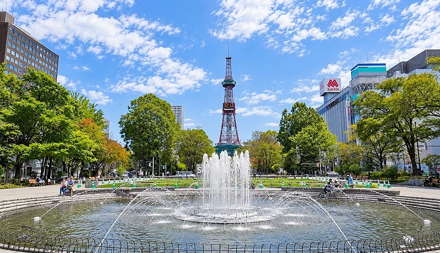 公園中央的噴水池後方有紅白色電視塔,兩側有綠樹與行人,右側大樓掛有「丸井今井」招牌,天空多雲