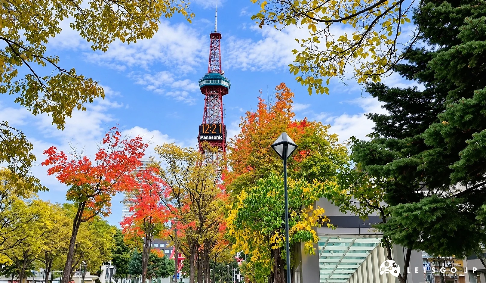 紅白色電視塔上有「Panasonic」電子時鐘,前方有紅黃色秋葉樹木與針葉樹,天空多雲