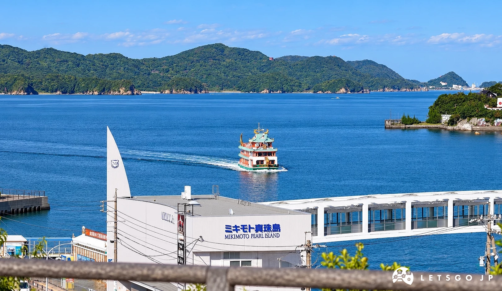 海面上有一艘多層紅綠色觀光船行駛,前方建築寫有「ミキモト真珠島」與「MIKIMOTO PEARL ISLAND」,遠處為綠色山丘。