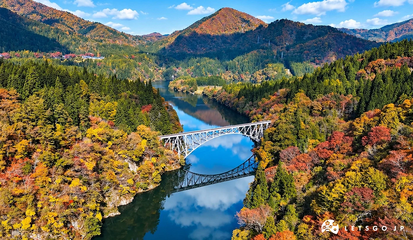 秋季山谷間的河面上橫跨一座鋼構拱橋,橋影與藍天白雲倒映水中,兩岸山林呈現紅黃綠色。