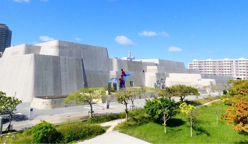 沖繩現代美術館建築,灰白混凝土幾何塊體外觀,前庭紅藍色抽象雕塑,綠色草坪與綠樹,藍天白雲,周邊白色公寓大樓,當代建築風格。