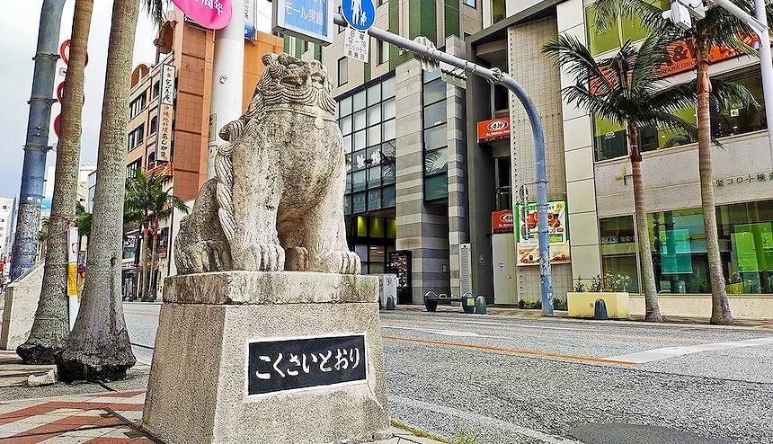 沖繩那霸國際通石獅雕像,灰白石材獅子座落於方形基座,黑底白字標示「こくさいどおり」,棕櫚樹與現代建築街景,晴朗陰天,南國城市風情。