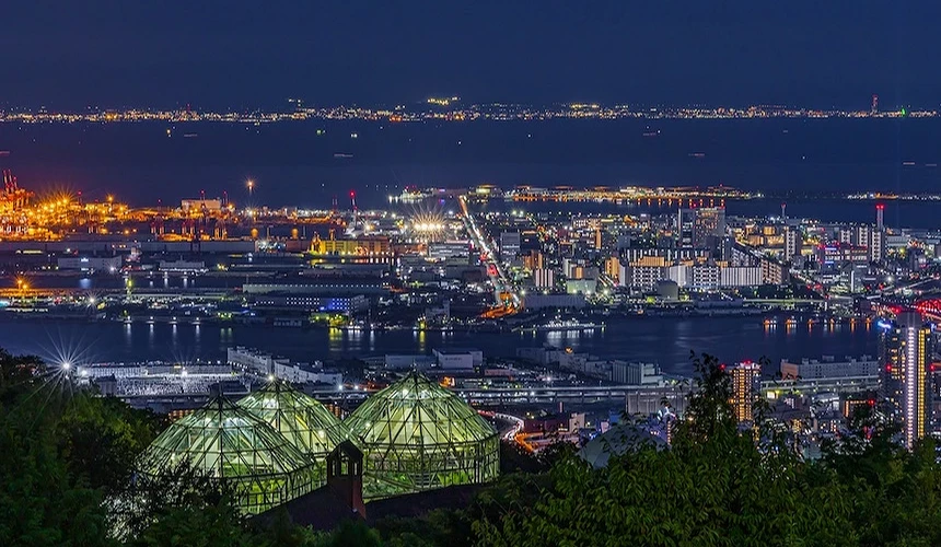山頂觀景設施夜景,綠色玻璃帳篷結構與黑色支架,俯瞰神戶港灣全景,金黃港口燈火與貨櫃堆場,遠景城市建築璀璨,深藍港灣與遠景山脈,夜間工業港景象壯觀。