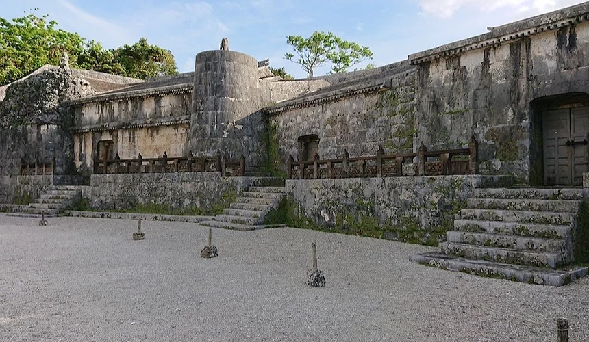 沖繩琉球石灰岩城郭遺跡,灰白石牆與圓形角樓,石階與木製欄杆,碎石廣場,綠樹與藍天白雲,古老城址風貌。