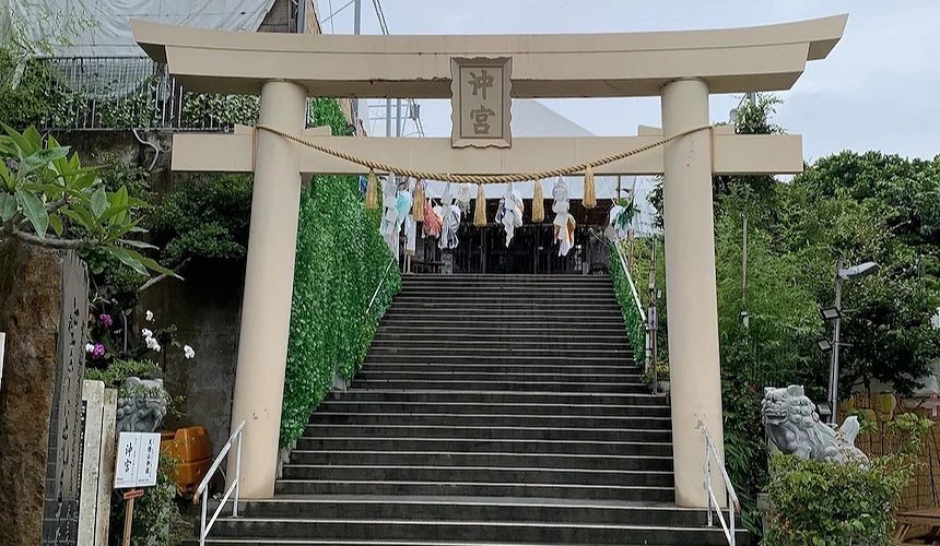 沖繩神社鳥居,米白色木構鳥居上掛「神宮」匾額,石階通往主殿,階梯兩側綠色植栽,石獅守護,懸掛白色紙垂與注連繩,陰天氛圍,南國神社風情。
