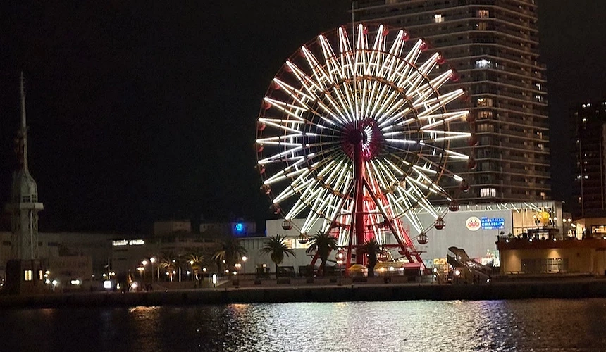 神戸摩天輪夜景,紅白色巨型輪轂與金色燈管,黑色夜空背景,高樓建築與棕櫚樹,平靜港灣水面映燈光,濱水娛樂設施璀璨。