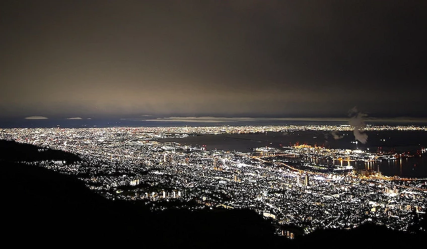 神戶港夜景全景,萬家燈火點綴城市,金黃燈光映照港灣與海面,遠景山脈輪廓,暗藍天空與烏雲,城市夜晚璀璨壯觀。