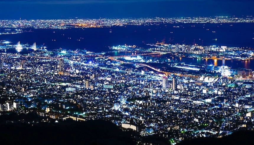 神戶城市夜景全景,萬家燈火鋪滿城市,藍綠色與金黃燈光交織,港灣與遠景山脈,深藍夜空,都市夜晚璀璨壯闊。