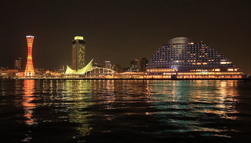 神戶港濱水夜景,橙紅塔樓與黃色高樓,白色帆形橋樑,紫色燈光大型飯店建築,金黃港灣燈火倒映水面,黑色夜空,港口建築群璀璨。