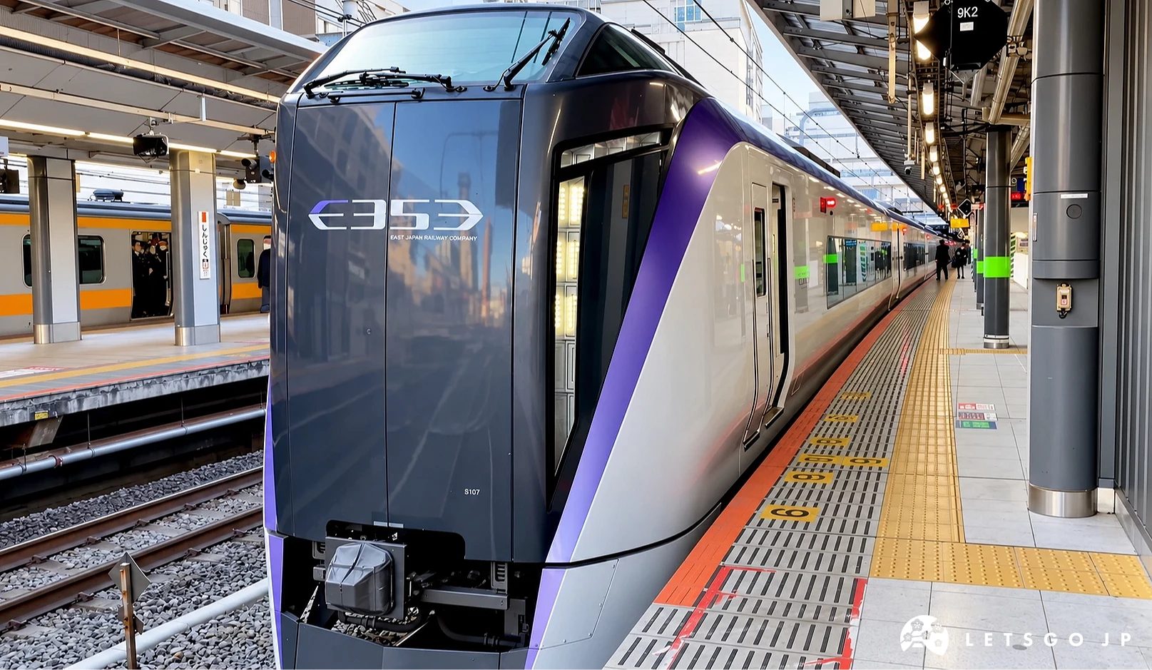 紫色線條的E353系特急列車停靠在月台,車頭印有E353標誌,旁邊軌道停有黃橘色電車