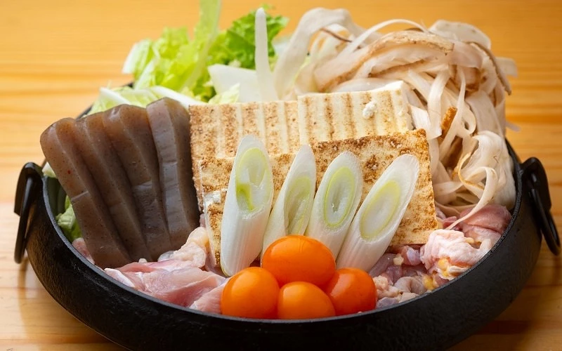 黑色鐵鍋內擺放蒟蒻、烤豆腐、蔥段、牛蒡片、生雞肉、蛋黃與白菜