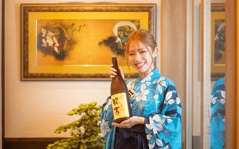 身穿藍色花紋浴衣的女子手持日本酒瓶微笑，背後有畫作與盆栽