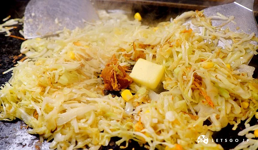 鐵板上攤炒文字燒食材,,中央置方形奶油塊,左右各有一把金屬鏟