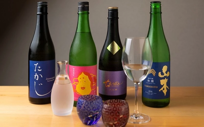四瓶日本酒與酒壺、兩個彩色酒杯、玻璃酒杯擺放在木桌上,酒標寫有「山本」