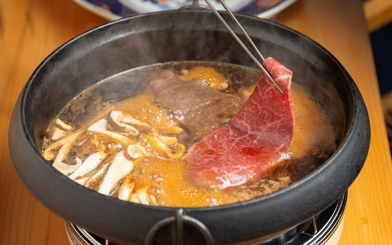 黑色鐵鍋在爐上煮著湯汁與菇類,筷子夾起一片生牛肉