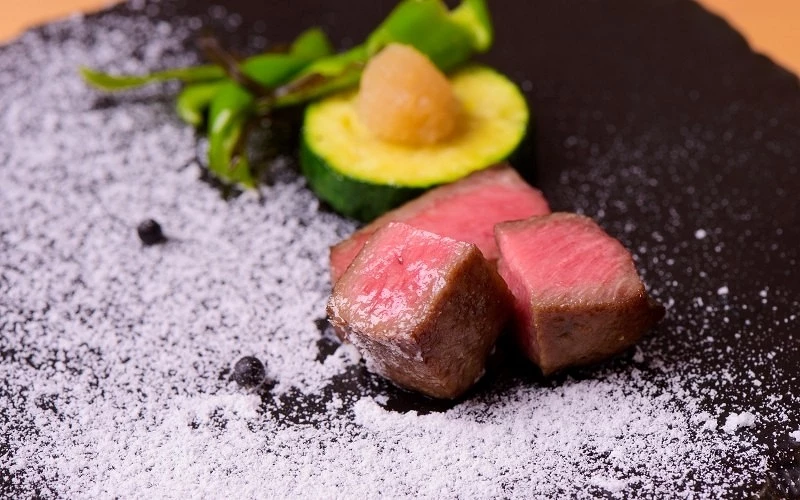 黑色石板上擺放兩塊牛肉塊,旁有櫛瓜切片、青椒與白色鹽粉