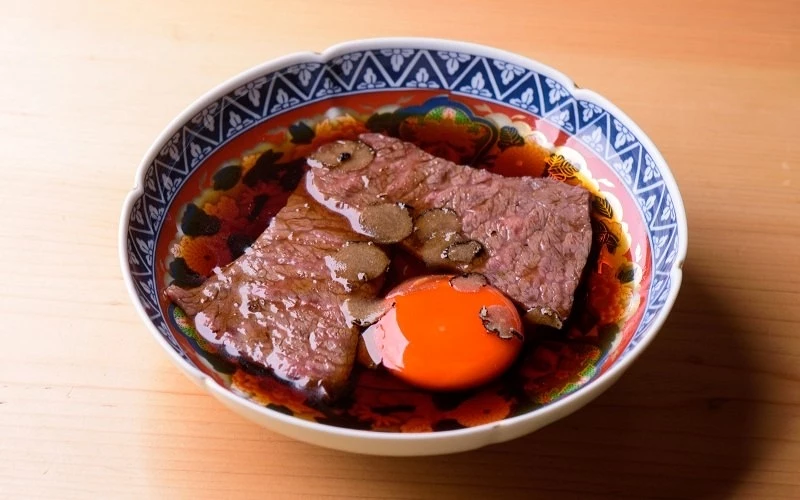 花紋碗中擺放兩片牛肉與黑松露片,旁有一顆蛋黃