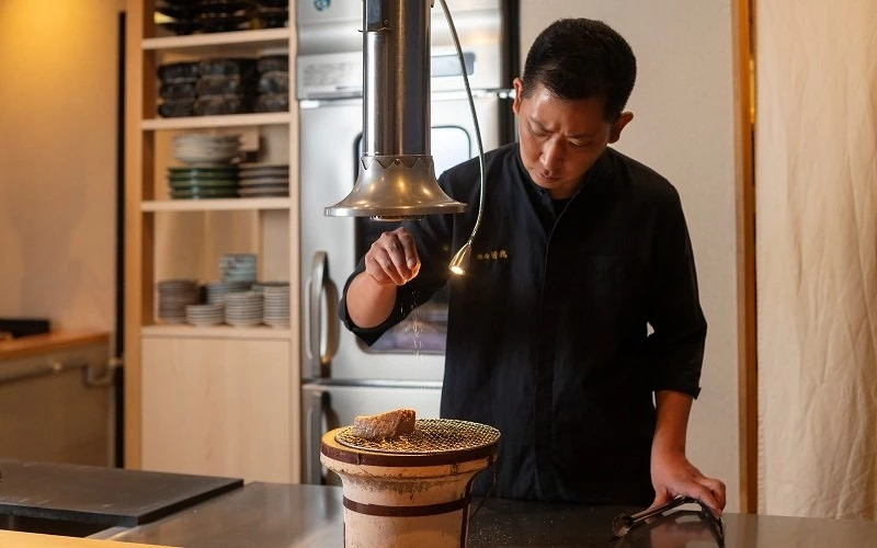 身穿黑色制服的廚師站在炭爐前,爐上網架放有一塊肉