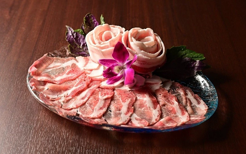 玻璃盤中盛放豬肉薄片，中央有兩朵豬肉捲與紫色蘭花、紫蘇葉
