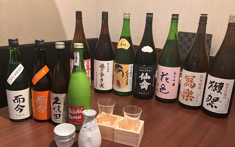 木桌上擺放多款日本清酒酒瓶，前方有酒壺、酒杯與木枡