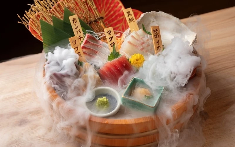 木桶中擺放插有品名木牌的生魚片拼盤,周圍冒出白色煙霧,旁附山葵與小菜