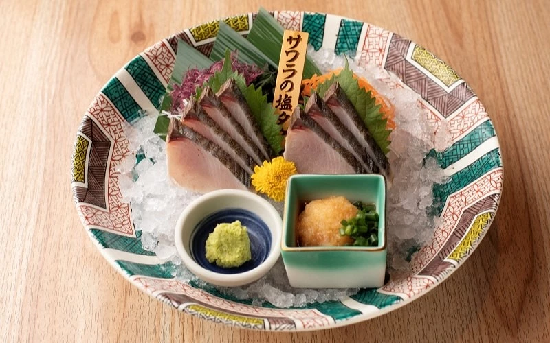花紋圓盤中的鰆魚生魚片鋪在碎冰上,木牌寫有「サワラの塩タ」,旁附山葵與蘿蔔泥