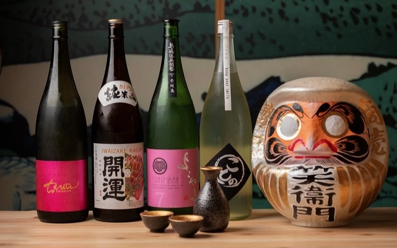 五瓶日本酒與酒壺、兩個酒杯擺放在木桌上,旁有寫有「笑衛門」的達摩