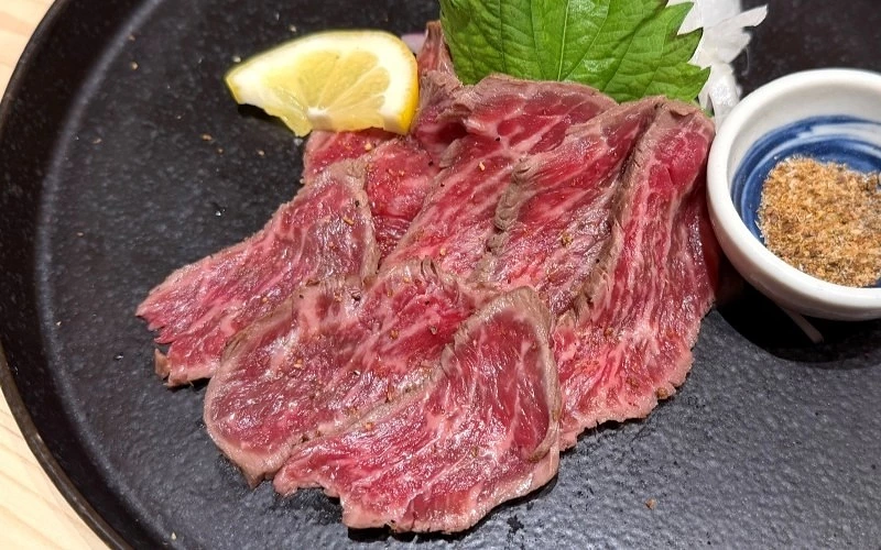 黑色圓盤上擺放數片切片生牛肉,旁附檸檬片、紫蘇葉與小碟調味粉
