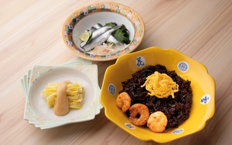 桌上擺放黑色炊飯配蝦、醃漬小魚與黃色蛋絲小菜三道料理