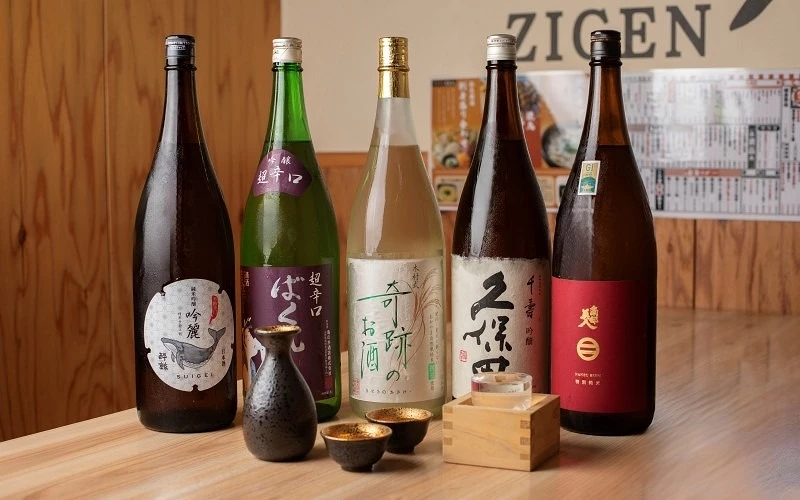 桌上擺放五瓶日本酒、酒壺、兩個酒杯與一個木升，背景牆面寫有 ZIGEN