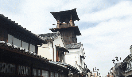 川越著名地標「時之鐘」木造鐘樓,周圍環繞著傳統黑瓦與白牆建築,背景為微雲的天空